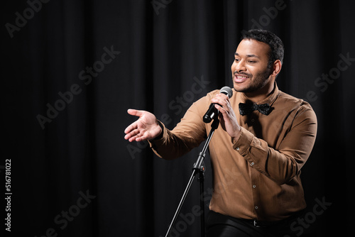indian comedian performing stand up comedy and telling jokes in microphone while gesturing on black.