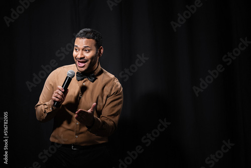 Valokuva positive indian comedian in shirt and bow tie holding microphone during monologue on black