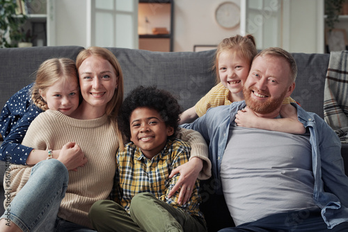 Portrait of big family with adoptive children embracing and smiling at camera sitting in living room at home