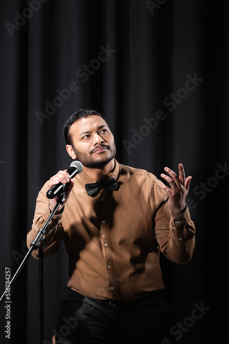 pensive indian comedian performing stand up comedy and telling jokes in microphone on black.