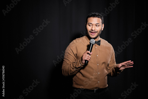 indian stand up comedian with bow tie holding microphone during monologue on black.