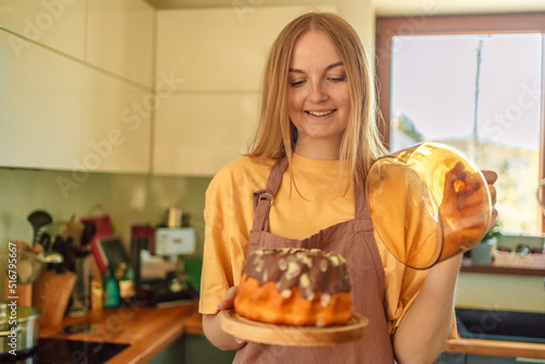 Smiling young blonde woman chef cook in apron standing at the kitchen, showing tasty pie. 