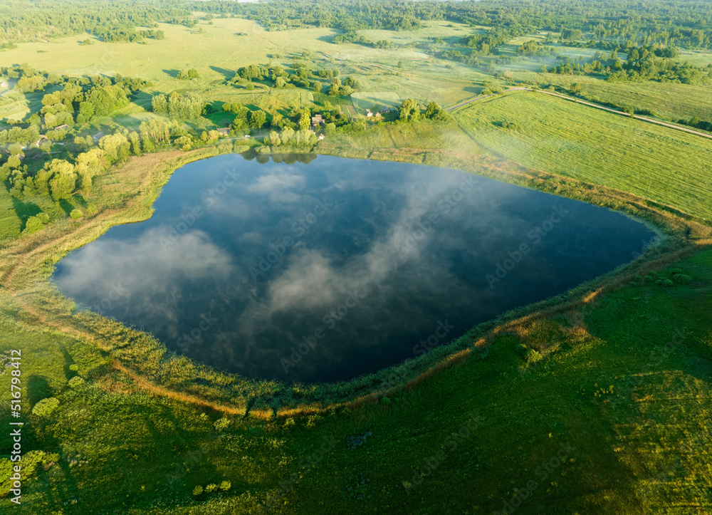 Fototapeta premium Foggy sunrise over lake. Nature at dawn in fog, aerial view. Morning mist haze on lake. Sunny morning scene in misty in rural. Misty landscape on pond.. Sunny foggy hills on sundown. Dawn in hillside.