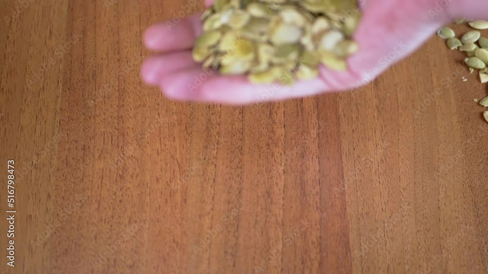 A Lot of Raw Pumpkin Seeds Fall from Female Hands on the Table. Thrown green pumpkin seeds fall through fingers into a pile of seeds on a desk. Scattered grains. Natural, nutritious, healthy product. A Lot of Raw Pumpkin Seeds Fall from Female Hands on the Table. Thrown green pumpkin seeds fall through fingers into a pile of seeds on a desk. Scattered grains. Natural, nutritious, healthy product.