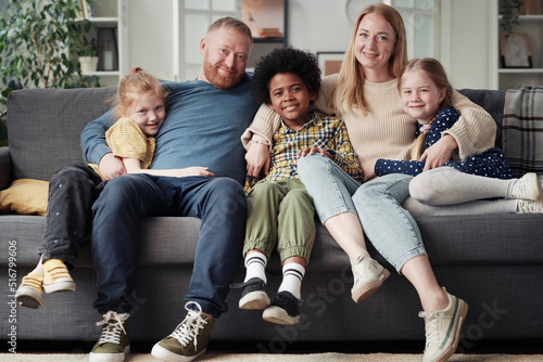 Portrait of big adoptive family with children sitting on sofa, embracing each other and smiling at camera