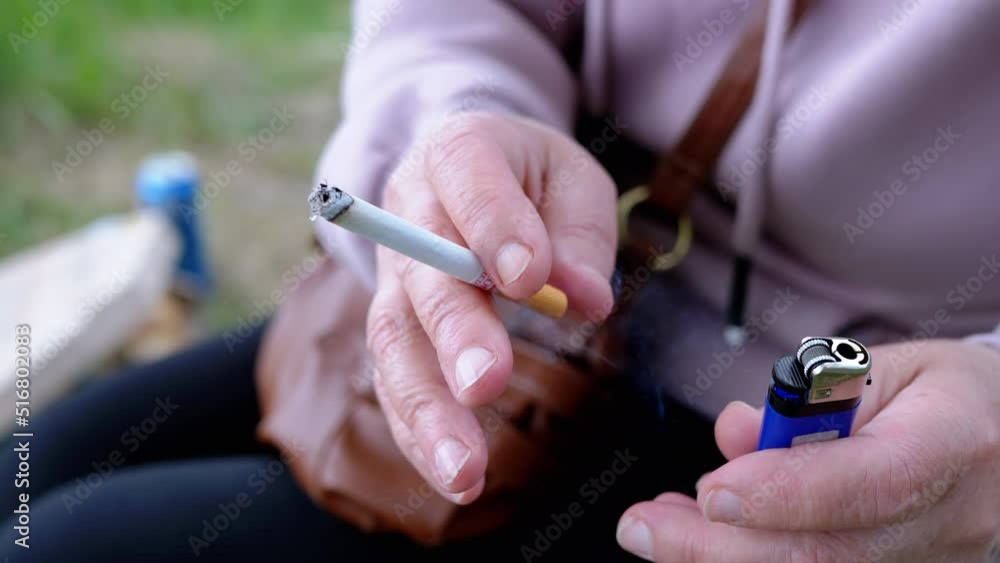 Woman Holding a Cigarette and a Lighter in Hands Sitting in Nature on Sun. A female smokes a fuming cigarette outdoors, park. Smoking. Bad habit. Addiction. Unhealthy lifestyles. Stress. Resting. Woman Holding a Cigarette and a Lighter in Hands Sitting in Nature on Sun. A female smokes a fuming cigarette outdoors, park. Smoking. Bad habit. Addiction. Unhealthy lifestyles. Stress. Resting.
