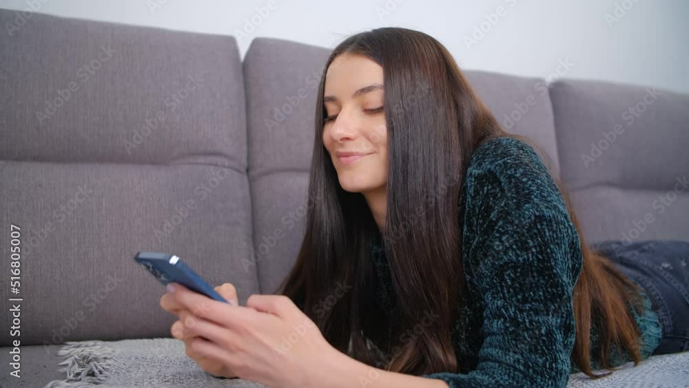 Young brunette woman with long dark hair lying on a couch at home and texting online with a cheerful smile. Happy female person in late 20s resting at home and typing a message on a smart phone