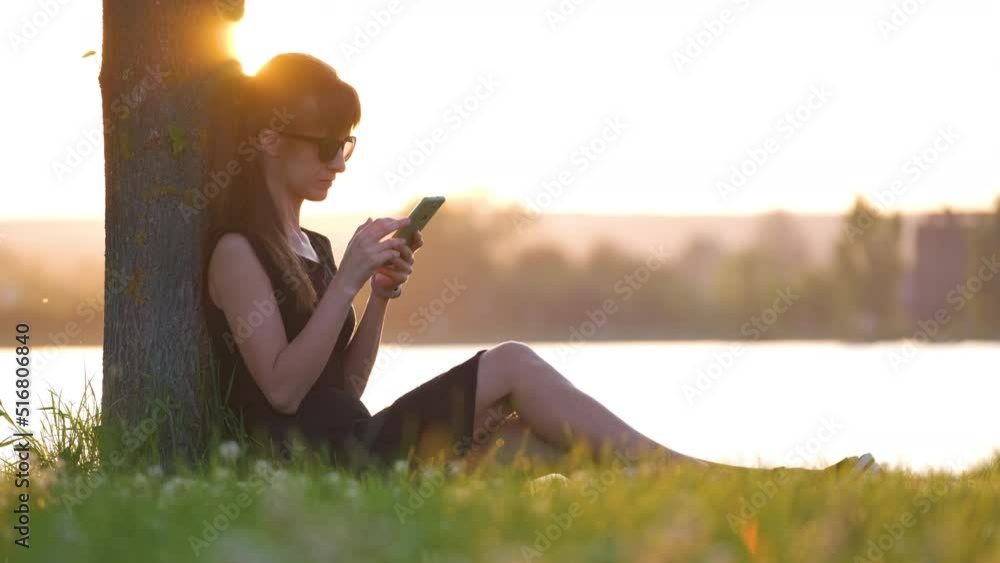 Young woman relaxing on lakeside grass lawn browsing her mobile phone outdoors on warm summer evening. Communication and mobile connection concept Young woman relaxing on lakeside grass lawn browsing her mobile phone outdoors on warm summer evening. Communication and mobile connection concept