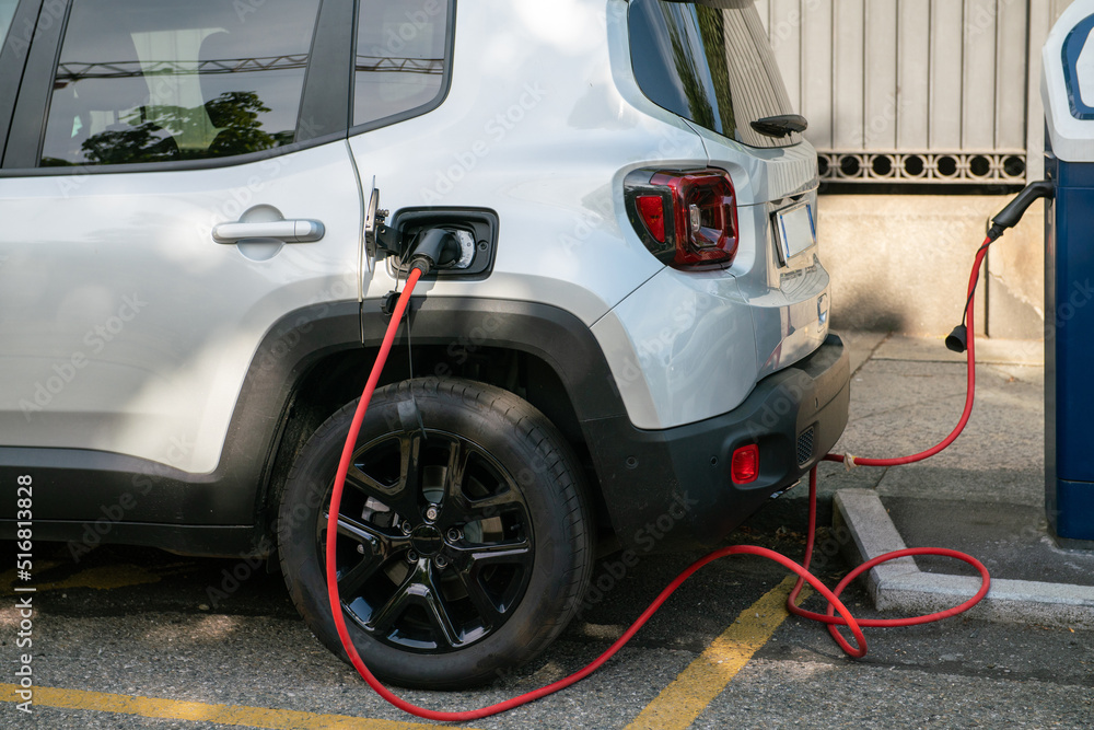 electric car being recharged, connected with the red cable to the electrical control unit, where it draws energy, in a dedicated parking lot. Zero emissions in cities with green ecological policies. electric car being recharged, connected with the red cable to the electrical control unit, where it draws energy, in a dedicated parking lot. Zero emissions in cities with green ecological policies.