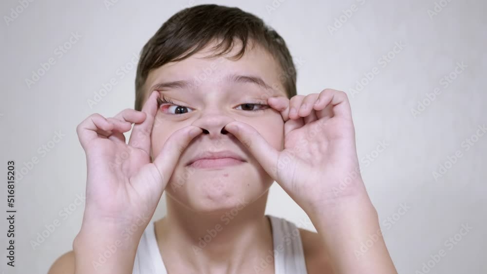 Positive Child Touches his Face with his Hands, Grimaces, Looking at the Camera. Boy makes funny faces, rolls back eyelids on eyes, opens mouth, spreads nostrils with his fingers. Funny antics. Humor. Positive Child Touches his Face with his Hands, Grimaces, Looking at the Camera. Boy makes funny faces, rolls back eyelids on eyes, opens mouth, spreads nostrils with his fingers. Funny antics. Humor.