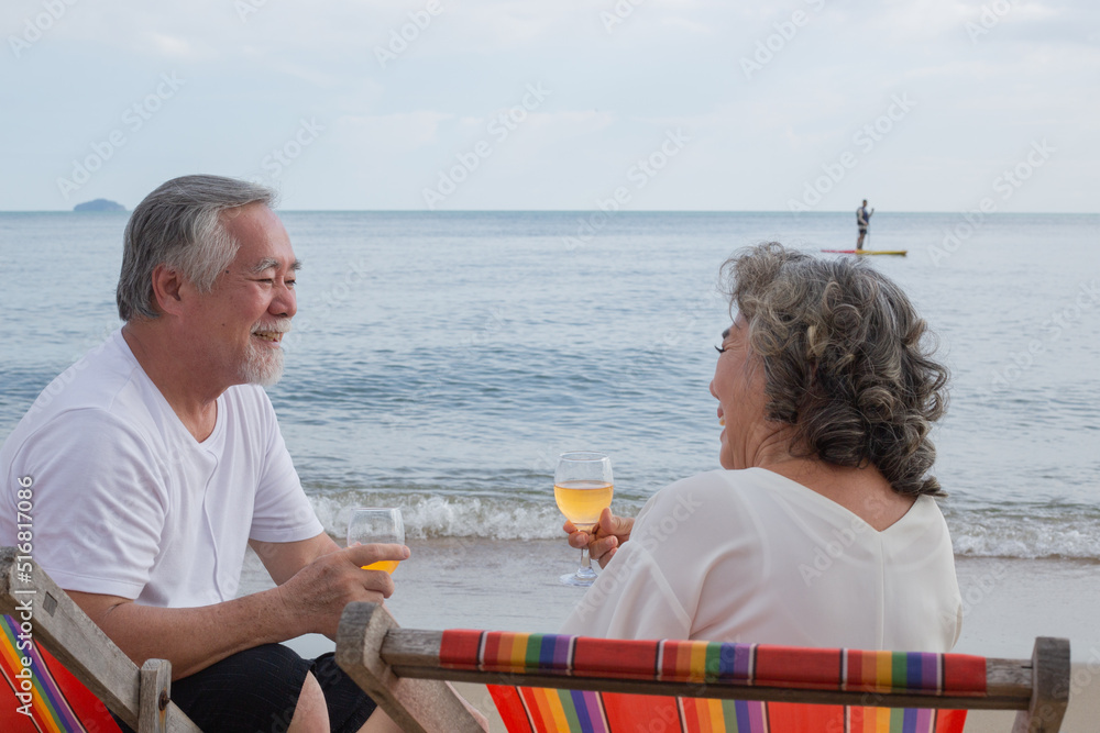 Asian senior couple toasting glasses and enjoy drinking wine, sitting and talking together at beach with sea and wave background. travel and vacation after retirement concept