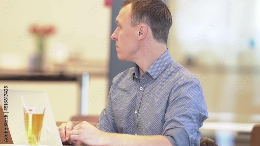 A young man sitting at a table with a laptop in a cafe looks away. A slow-motion average plan of a man. The concept of remote work. High quality FullHD footage A young man sitting at a table with a laptop in a cafe looks away. A slow-motion average plan of a man. The concept of remote work. High quality FullHD footage