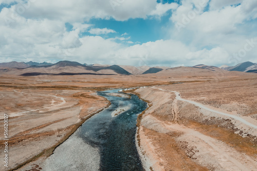 blue natural river flowing between dry desolate plateau with mountain range on horizon in Deosai National Park in Pakistan, aerial