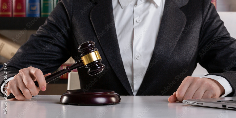 Female judge hands in formal wear with maroon auction gavel and open laptop on a desk surface in an office, front view, selected focus.