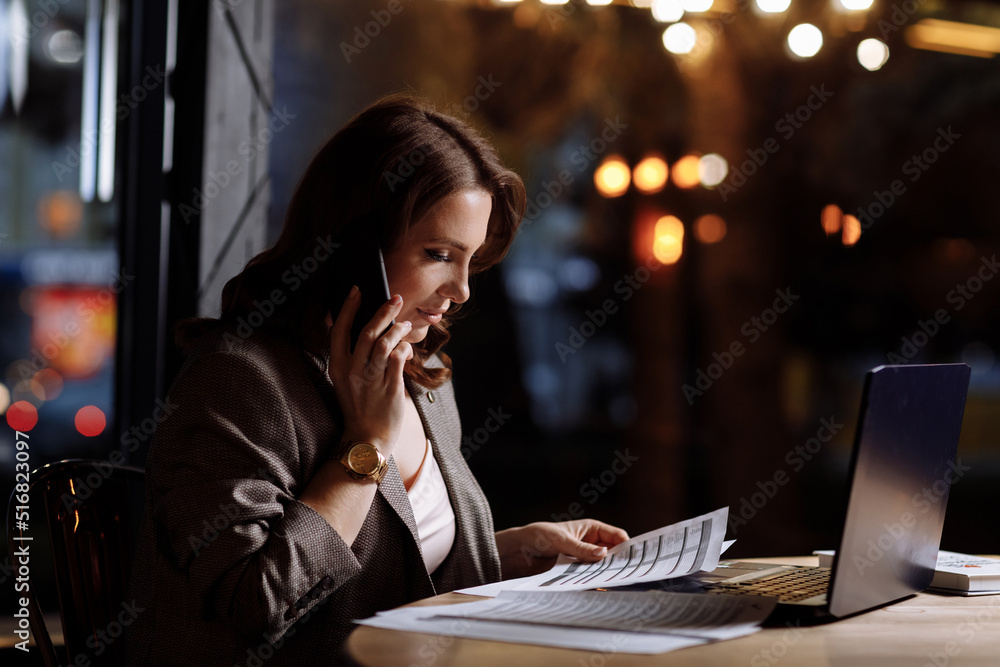 smiling successful beautiful young businesswoman working in cafe. female is talking cellphone while working remotely in elegant wooden interior with many greens. laptop with documents on table. smiling successful beautiful young businesswoman working in cafe. female is talking cellphone while working remotely in elegant wooden interior with many greens. laptop with documents on table.