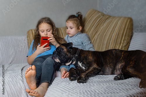 two little sister girls are sitting at home on the couch and watching the phone, with them their four-legged friend dog breed German boxer sleeps peacefully