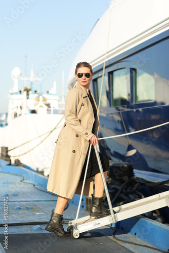 young slavic woman in sun glasses with long hair wearing trench coat boarding on ship for traveling full body photo
