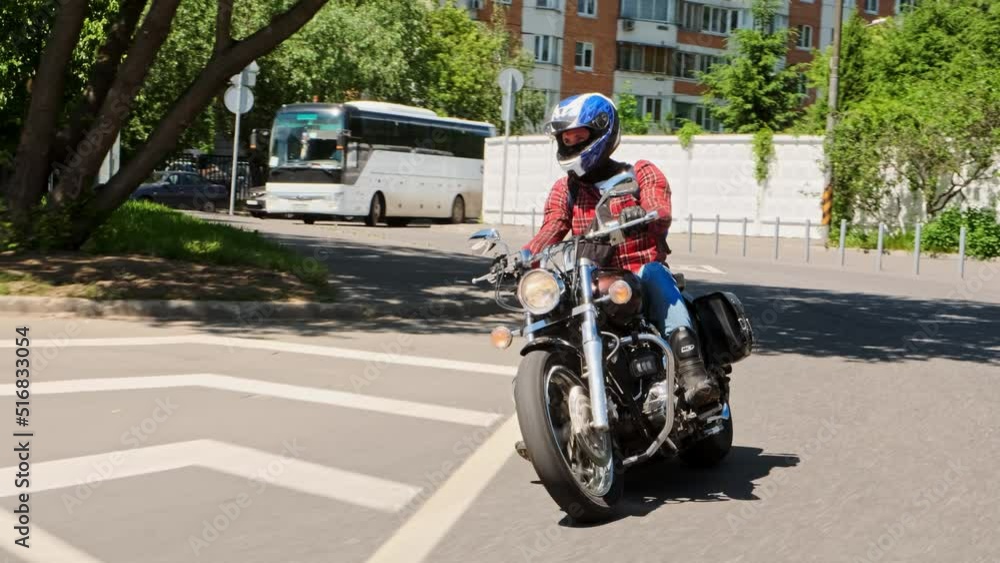 Front view biker in red shirt and blue helmet in jeans drives his brown dragster down road through city with trees on background. man rides motorcycle or bike on city street near park.
