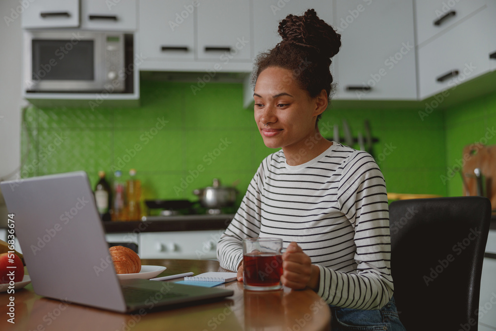 Beautiful smiling young woman holding cup of hot drink using laptop sitting at home Beautiful smiling young woman holding cup of hot drink using laptop sitting at home