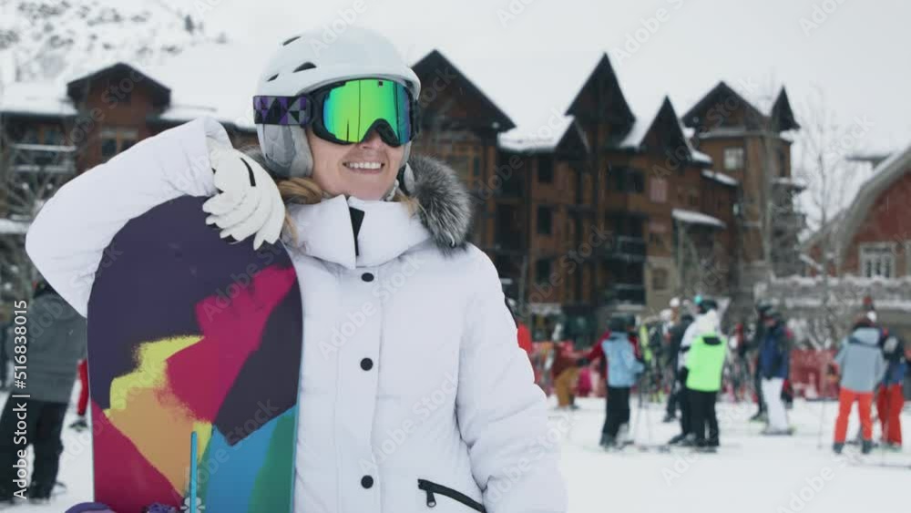 Woman enjoying leisure time after riding down mountain on snowboard. Female in snowboarder outwear googles with helmet waiving hand to her friends with winter chalet in ski resort on blurry background Woman enjoying leisure time after riding down mountain on snowboard. Female in snowboarder outwear googles with helmet waiving hand to her friends with winter chalet in ski resort on blurry background