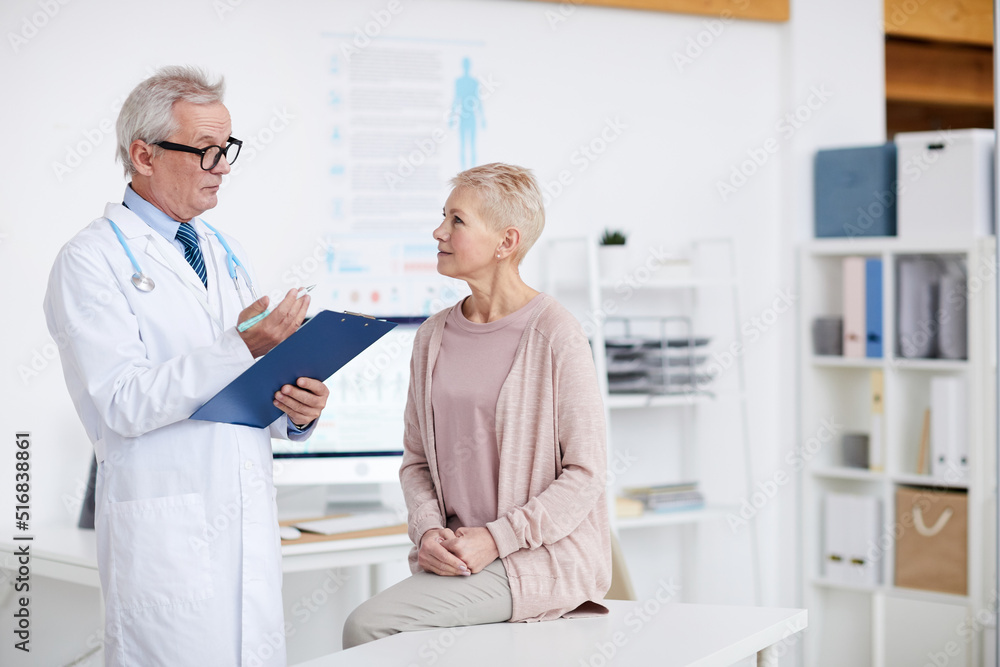 Serious attractive senior woman sitting on examination table and listening to qualified doctor at appointment Serious attractive senior woman sitting on examination table and listening to qualified doctor at appointment
