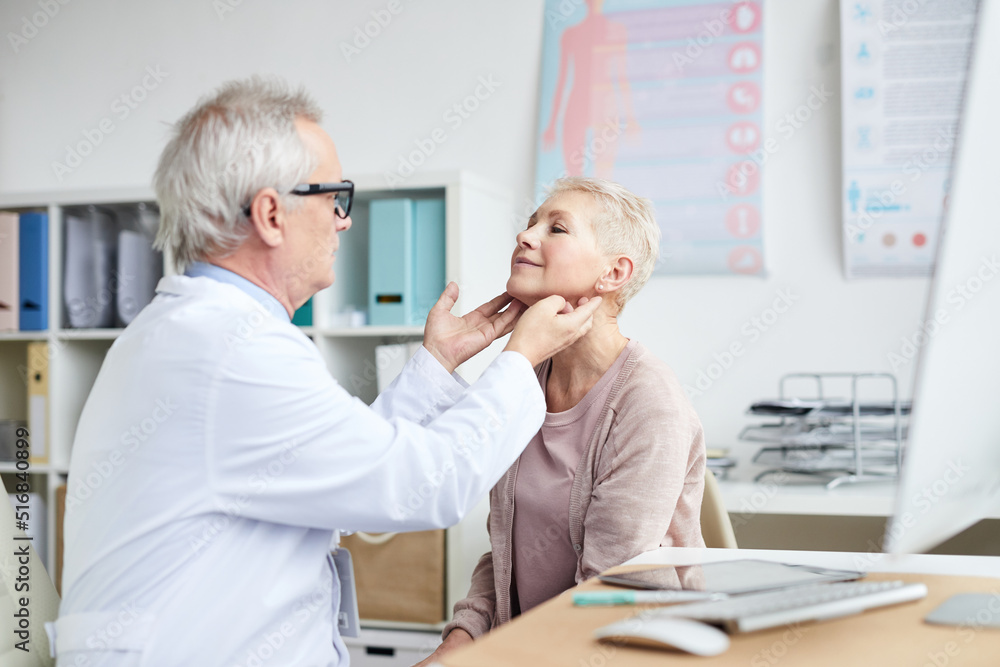 Obraz premium Serious senior male oncologist with gray hair sitting in front of mature female patient and touching her neck during checkups
