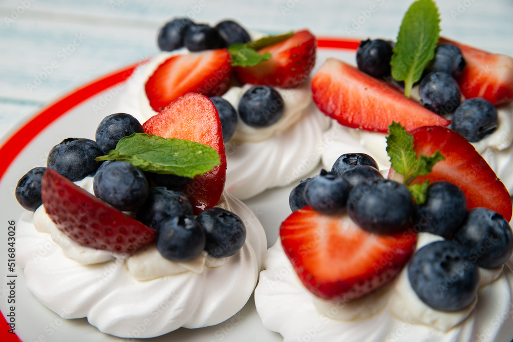 Four cakes Anna Pavlova with blueberries and strawberries with a mint leaf on a beautiful plate. Home confectionery. Old retro white and blue table. Four cakes Anna Pavlova with blueberries and strawberries with a mint leaf on a beautiful plate. Home confectionery. Old retro white and blue table.
