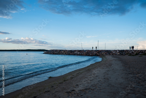 People silhouettes walking on a hard rock pier during summer sunset. Summer evening in a small village port.