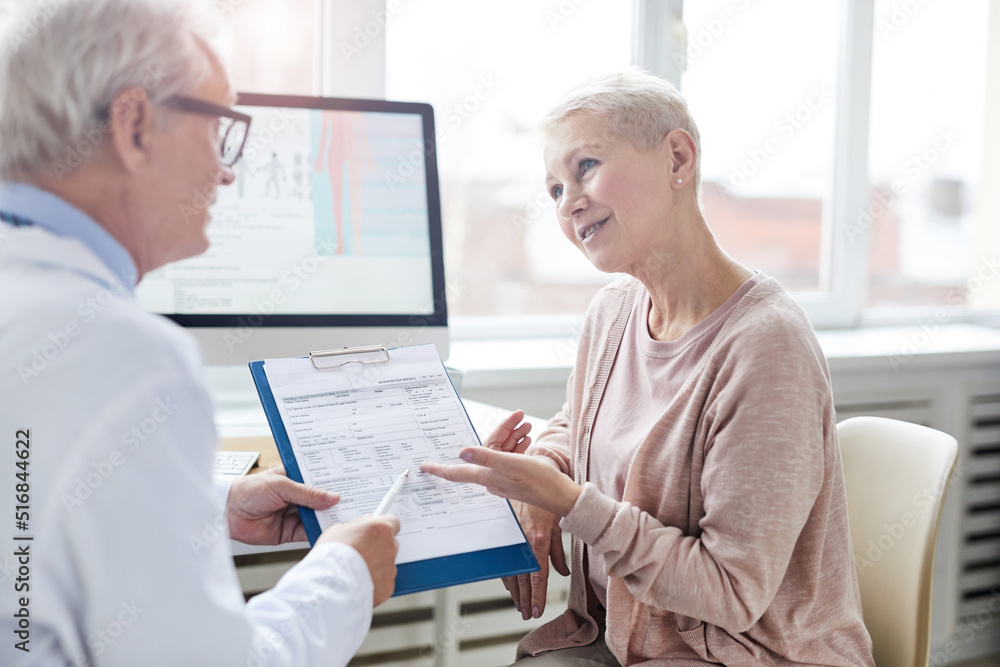 Obraz premium Smiling senior female patient with blond hair sitting at table and pointing at paper in clipboard while asking doctor about prescribed drugs