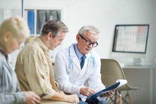 Serious aged male doctor with stethoscope around neck reading medical history of patient while holding consultation