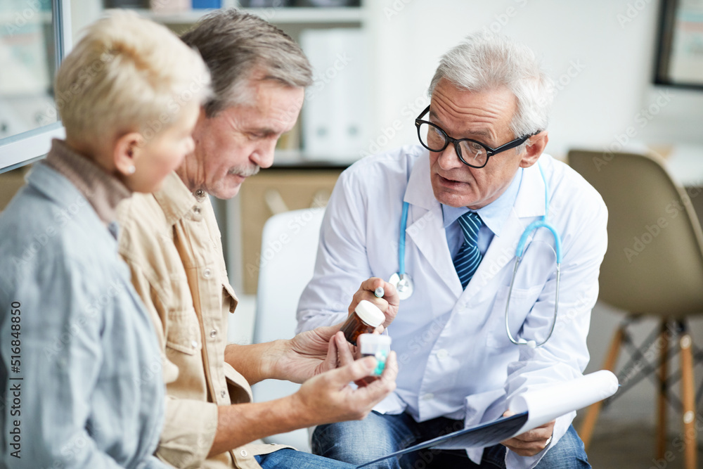 Obraz premium Confident male doctor in white coat pointing at pill bottle and recommending medicine to senior couple