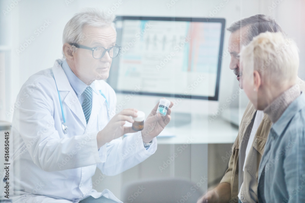 Behind glass view of senior doctor in eyeglasses holding pill bottles and explaining patients how to take pill Behind glass view of senior doctor in eyeglasses holding pill bottles and explaining patients how to take pill