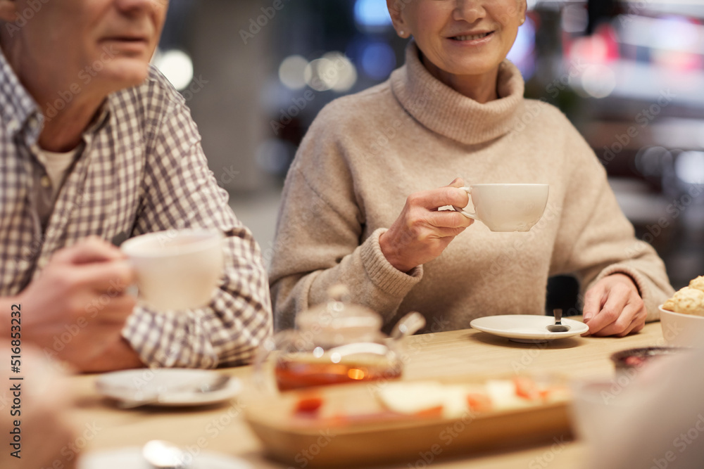 Close-up of positive seniors in casual clothing sitting at table and drinking tea while discussing last news in cafe