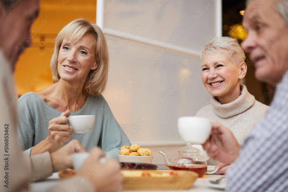 Group of smiling carefree mature friends sitting at table at home and chatting over tea cup