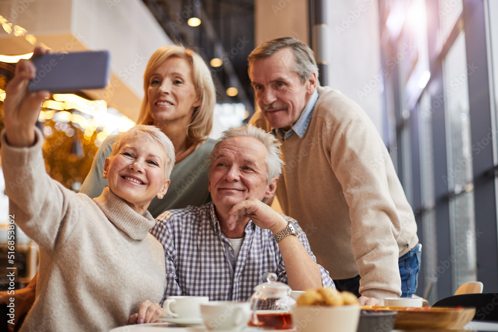Group of smiling senior friends in casual outfits using smartphone while taking selfie together in cafe Group of smiling senior friends in casual outfits using smartphone while taking selfie together in cafe