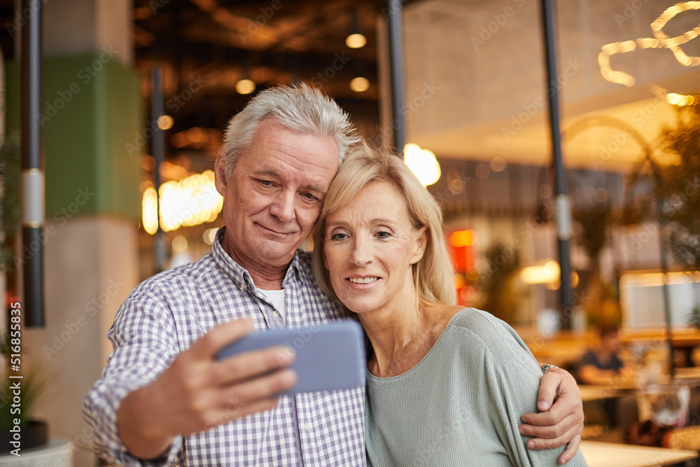 Handsome senior man with wrinkles embracing wife while posing with her for selfie on phone