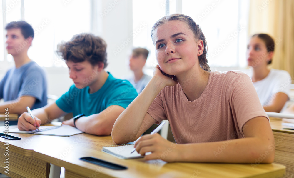 Obraz premium Portrait of cute dreamy smiling teenage girl sitting at school desk during lesson, resting her head on her hand
