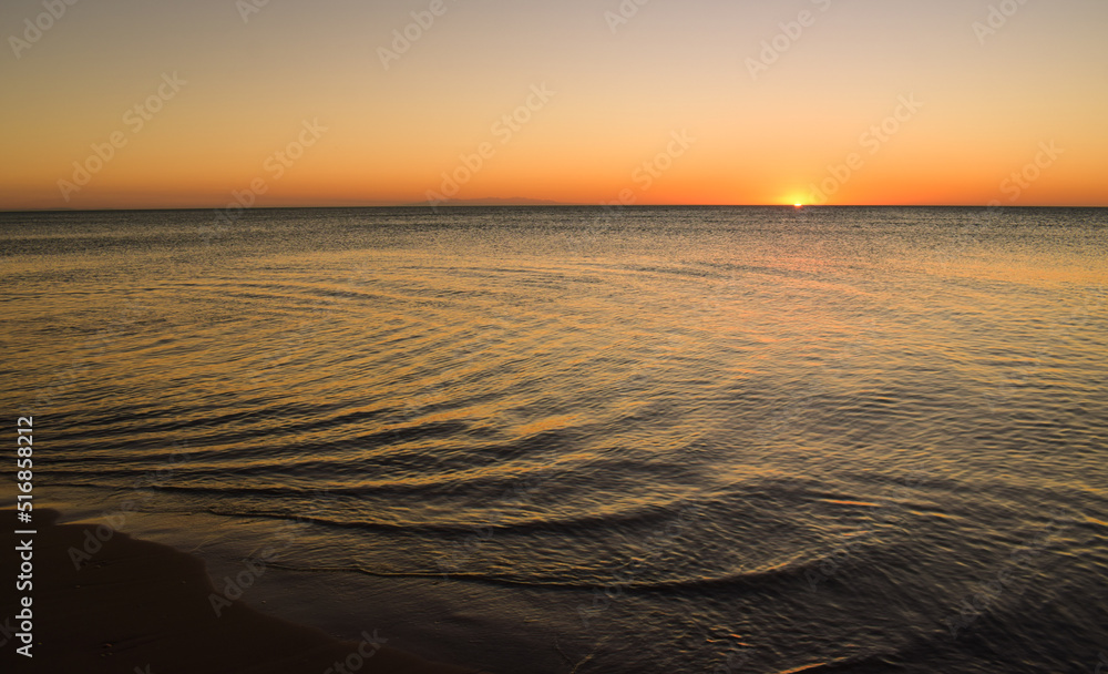 Sunset over the sea in summer time. The orange and yellow of the sky are reflected on the beach the while the sun comes down
