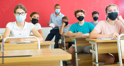 Young students in masks sitting in classroom and listening to male teacher.