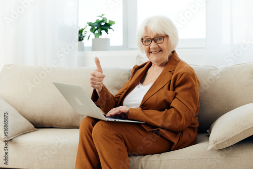 a joyful elderly woman is sitting on the sofa in a bright apartment in a brown suit, holding a laptop on her lap and pleasantly smiling with happiness showing a hand gesture as a sign of success