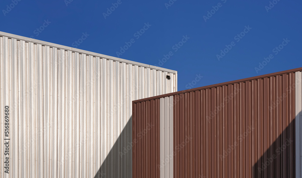 Sunlight and shadow on surface of 2 brown and silver corrugated steel warehouse against blue clear sky background