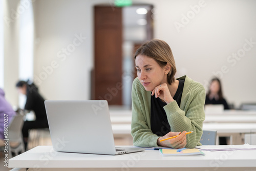 Online learning for adults. Pensive middle-aged woman studying on laptop in auditorium, using virtual learning platform. Mature female student engaged in study, watching webinar or attending e-class
