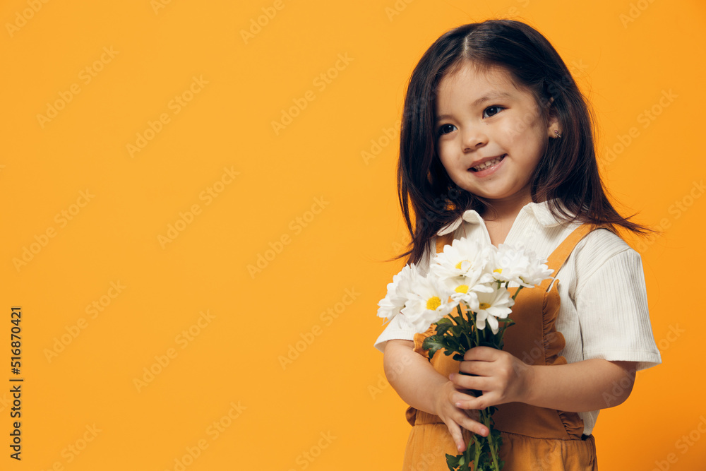 an adorable, attractive little girl in an orange sundress stands with a bouquet of daisies in her hand and smiles joyfully looking at the camera. an adorable, attractive little girl in an orange sundress stands with a bouquet of daisies in her hand and smiles joyfully looking at the camera.