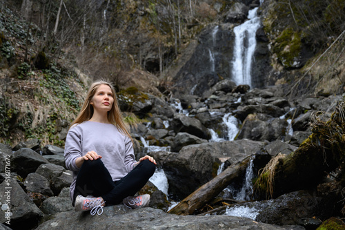 Girl sits on a large stone and meditates near a waterfall next to a mountain river.