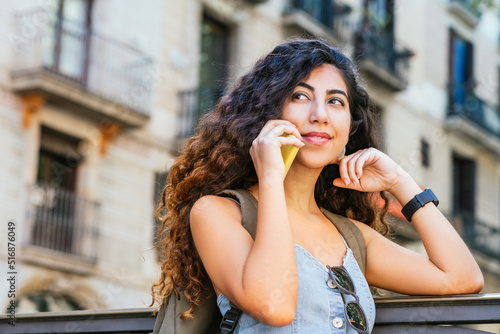 Cheerful indian woman with backpack answering phone call and looking away. She is leaning on a fence in the streets of Barcelona old town