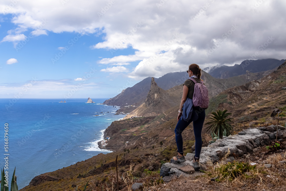 Backpack woman with scenic view of Atlantic Ocean coastline and Anaga mountain range on Tenerife, Canary Islands, Spain, Europe. Looking at Roque de las Animas crag. Hiking trail from Afur to Taganana