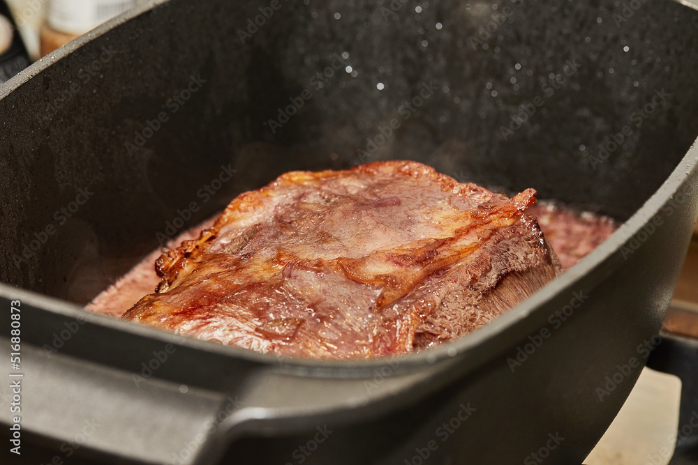 Piece of meat is fried with onions in wine sauce in rectangular pan over fire on gas stove
