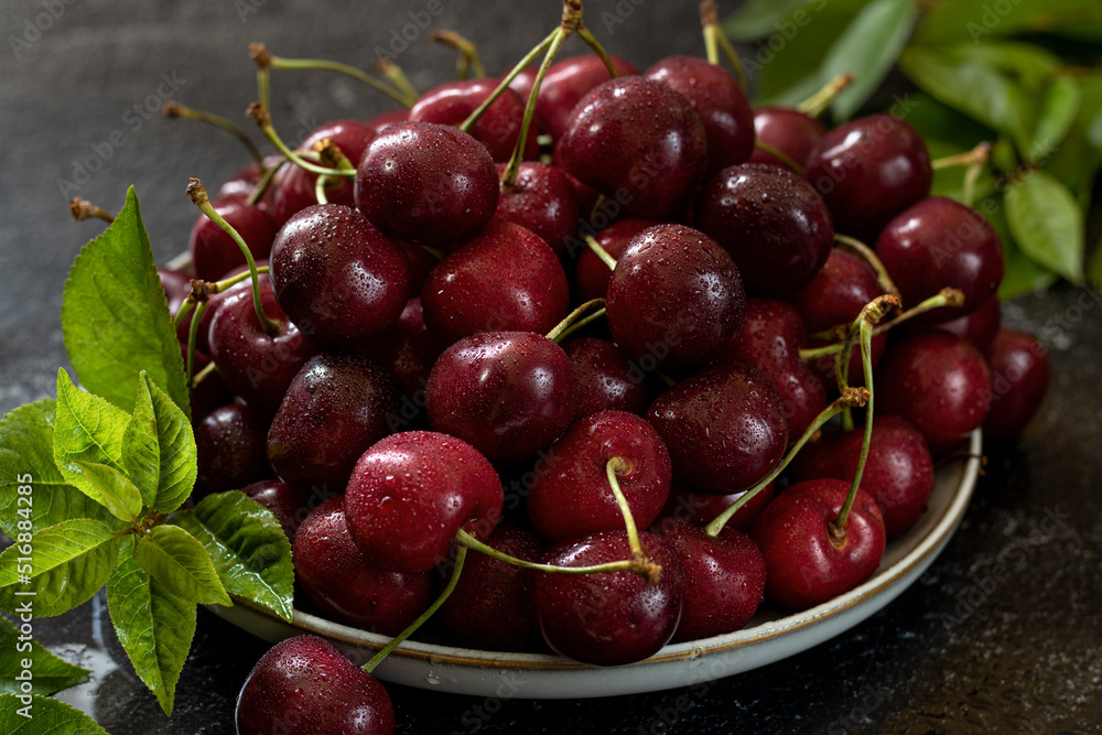 White ceramic dish filled with cherries, cherry leaves diagonally. Dark background. White ceramic dish filled with cherries, cherry leaves diagonally. Dark background.