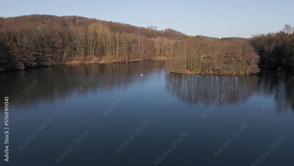 Big blue lake and leafless brown trees on its shores during a vibrant sunset in winter. Low flyover drone shot underneath a clear blue sky Big blue lake and leafless brown trees on its shores during a vibrant sunset in winter. Low flyover drone shot underneath a clear blue sky