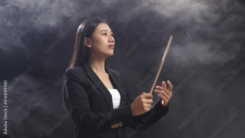 Side View Of Asian Conductor Woman Holding A Baton And Showing Gesture Quickly In The Black Studio With Fog 
